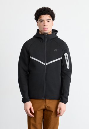 Sort zip-up hættetrøje med høj krave, der har reflekterende detaljer og et logo på brystet. Sammen med brune bukser.