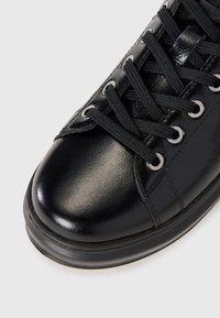 Black glossy leather sneaker with black laces threaded through metal eyelets, shown from a close-up top angle.