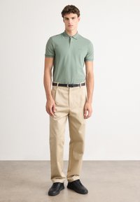 Grønn polo shirt med krage, beige skridde bukser og svarte sneakers som står på en jevn overflate. Enkel, klassisk design med minimal detaljer.