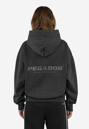 Person med brunt hår iført en sort hættetrøje med teksten "PEGADOR" på ryggen og sorte bukser med et lille "P" logo.