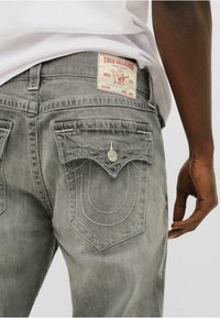 Jeans din denim gri, cu un etichetă semnătură True Religion la talie și cu buzunare din spate duble arcuitoare distinctive, cu cusături albe.