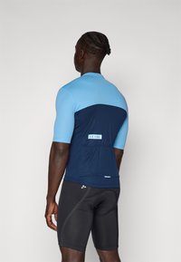 Maillot de cyclisme pour homme en bleu clair et marine, présentant un design ajusté, des manches courtes et une poche à l'arrière. Le matériau semble léger et respirant.