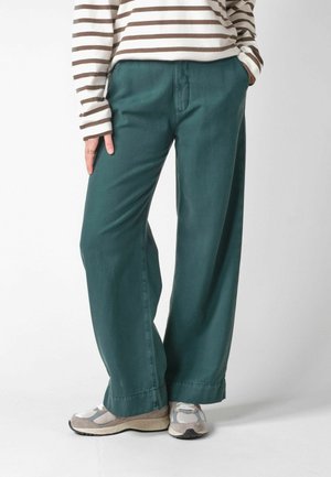 Personne portant un pantalon large couleur sarcelle, un t-shirt à manches longues rayé et des baskets blanches et grises, debout avec les mains dans les poches.