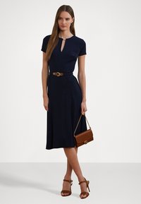 Abito blu navy con maniche corte e scollatura a chiave, stretto in vita con una cintura marrone. Abbinato a una borsa marrone e tacchi marroni.