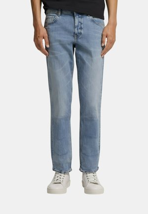 Uomo che indossa jeans a gamba dritta azzurri chiaro e sneakers bianche con lacci, in piedi contro uno sfondo neutro.