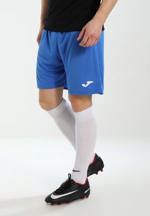 Persoon draagt blauwe sportshorts, witte kniekousen en zwarte voetbalschoenen, staand op een witte achtergrond.