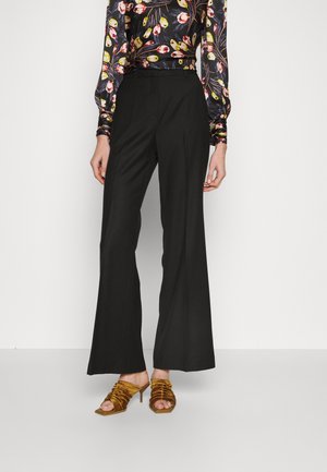 Kobieta ubrana w czarne spodnie z szerokimi nogawkami, kwiatową bluzkę z długim rękawem oraz złote marszczone sandały na wysokim obcasie, stojąca na białym tle.