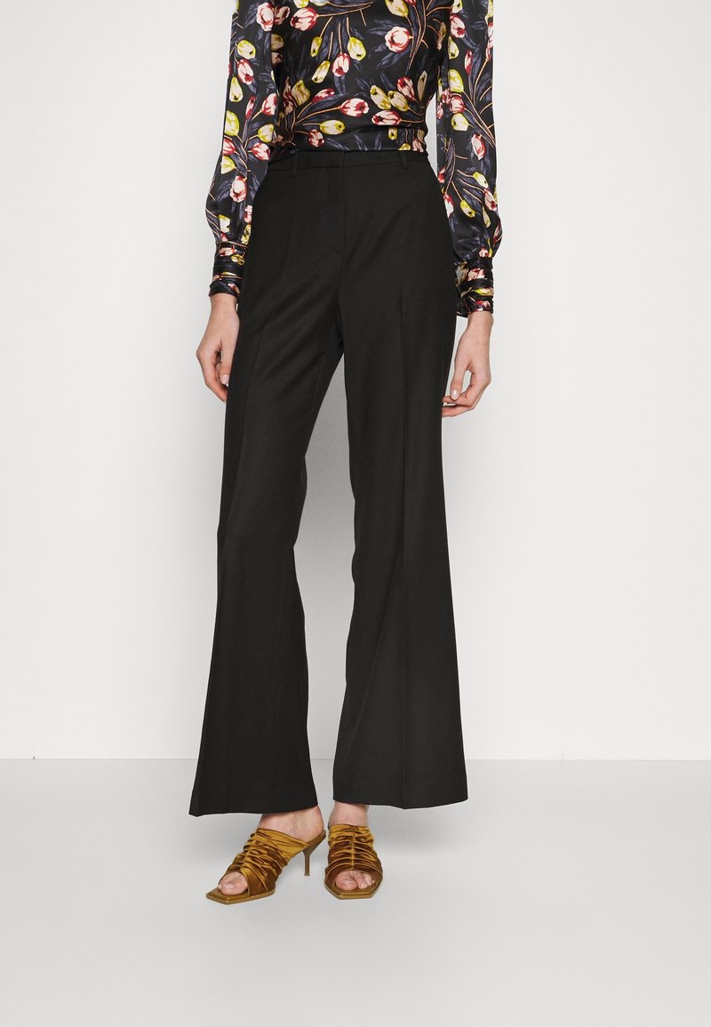 Femme portant un pantalon noir à jambes larges, un chemisier fleuri à manches longues et des sandales dorées à talons hauts plissées, debout sur fond blanc.