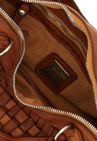 Interno di borsa in pelle marrone con texture intrecciata, fodera color cuoio, cerniera argentata e un'etichetta con testo in rilievo che dettaglia il marchio e la data.