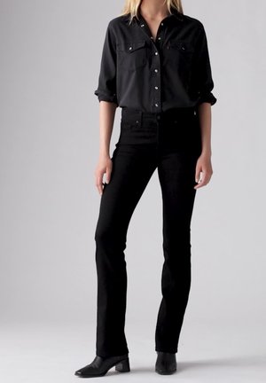 Kvinde iført en sort skjorte med knapper og op rullede ærmer, tætsiddende sorte bukser og sorte ankelstøvler, der står mod en ensfarvet baggrund.