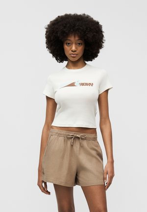 Młoda kobieta o afro fryzurze, ubraną w białą skróconą koszulkę z grafiką oraz beżowe szorty z wiązaniem, stojąca na jednolitym tle.