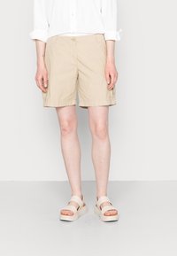 Donna che indossa pantaloni corti beige al ginocchio, una camicia bianca a maniche lunghe con maniche arrotolate e sandali con piattaforma beige, in piedi su uno sfondo neutro.