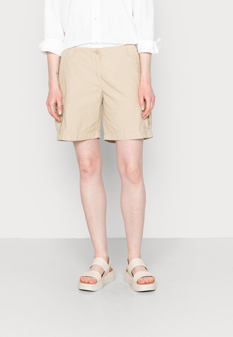 Donna che indossa pantaloni corti beige al ginocchio, una camicia bianca a maniche lunghe con maniche arrotolate e sandali con piattaforma beige, in piedi su uno sfondo neutro.