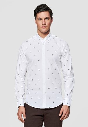 Mężczyzna ubrany w białą koszulę na guziki z długim rękawem, ozdobioną drobnym czarnym kwiatowym wzorem, oraz ciemne spodnie, stojący na jednolitym tle.