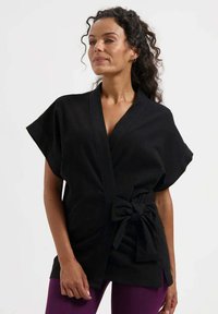 Schwarzes Oberteil im Kimono-Stil mit weiten Ärmeln, Taillenband und seitlichen Taschen. Glatter Stoff, minimalistisches Design, geeignet für den Freizeitgebrauch.