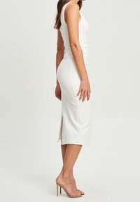 Bílé bezrukávové šaty z hladkého materiálu, přiléhavý střih, ke kolenům s bočním rozparkem, doplněné o nahé vysoké podpatky.