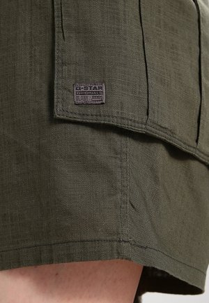 Nærbillede af olivengrønne stofshorts med en lille rektangulær lap med teksten G-Star Originals Raw Denim på lommelåget.