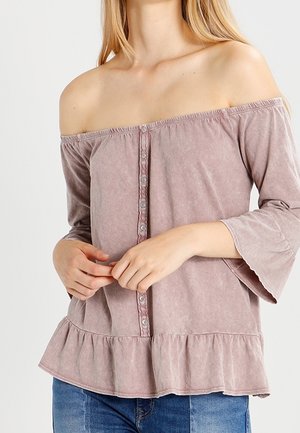 Femme portant un chemisier rose poussière à épaules dénudées avec des boutons à l'avant, des manches évasées et un ourlet volanté, assorti à un jean bleu.