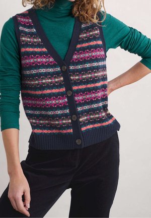 Kvinde med krøllet hår iført en mørkeblå knappet vest med flerfarvede mønstre over en grøn langærmet rullekrave og sorte bukser.