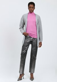 Helle graue Strickjacke, pinkfarbener Rollkragenpullover, metallisch silberne Hose und spitze schwarze Absätze mit Verzierungen. Glatte Texturen und figurbetontes Design.