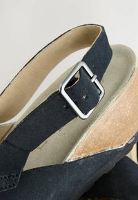 Sandale en suède noir avec une bride pour la cheville, ornée d'une boucle carrée en argent et d'une semelle en liège naturel. Doublure beige douce visible.