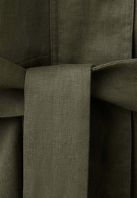 Detailný záber olivovo zeleného látkového opasku zviazaného uzlom okolo zodpovedajúceho textúrovaného ľanového materiálu.