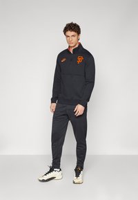 Sudadera negra de media cremallera con el logo naranja de San Francisco y el swoosh de Nike, combinada con joggers negros a juego y zapatillas deportivas color crema.