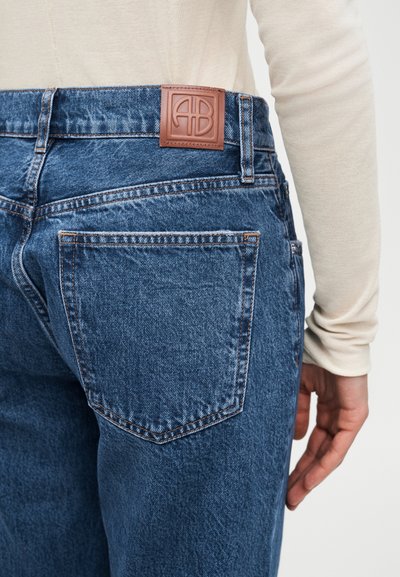 Person wearing blue denim jeans with a brown leather logo patch on the waistband, standing with their right arm relaxed by their side.