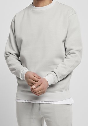 Mann trägt hellgrauen Pullover und passende Jogginghose, stellt seine Hände vorne ein, mit einem weißen Hemd darunter.