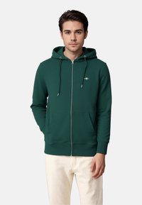 Grøn zip-up hættejakke lavet af blødt stof; har en frontlynlås, justerbare snore og en frontlomme. Klassisk afslappet design.