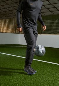 Schwarzes Nike Trainingstop und -hose, schwarze Fußballschuhe mit blauen Akzenten, dribbelt einen grauen Fußball auf grünem Kunstrasen.