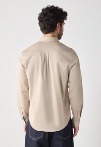 Camicia beige a maniche lunghe con colletto classico, piega posteriore e polsini con bottoni. Orlo arrotondato e tessuto morbido. Indossata con denim scuro.