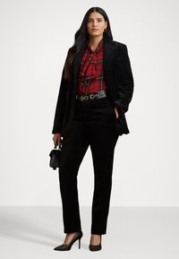 Giacca e pantaloni in velluto nero abbinati a una blusa a quadri rossi con cravatta a fiocco. Cintura in pelle nera e scarpe nere con tacco alto. Piccola borsa nera.