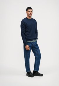 Navy sweatshirt with long sleeves over a green shirt, paired with blue denim jeans and black shoes. Simple design with a relaxed fit.