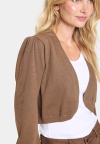 Brun kort cardigan i ett ribbat trikåtyg, med puffärmar och en djup V-ringning, lagd över en vit topp.