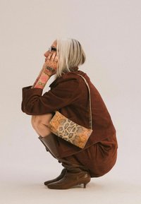 Bruine suède jas en jurk, gemêleerde bruine kniehoge laarzen en een crossbody tas met luipaardprint. Zilverkleurig haar, zichtbare tatoeages.