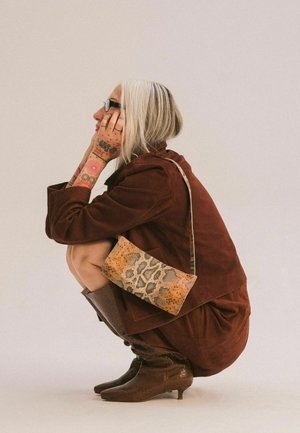 Bruine suède jas en jurk, gemêleerde bruine kniehoge laarzen en een crossbody tas met luipaardprint. Zilverkleurig haar, zichtbare tatoeages.