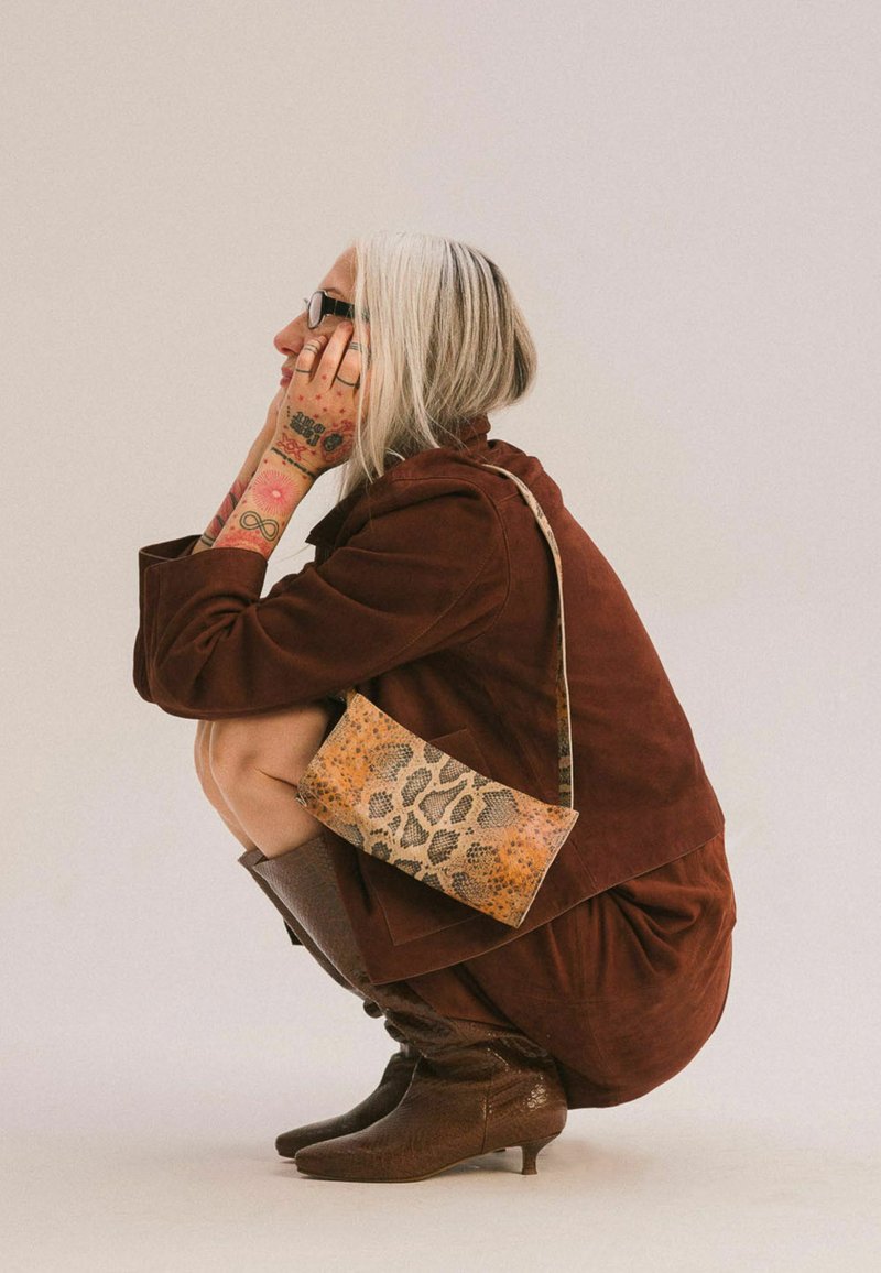 Bruine suède jas en jurk, gemêleerde bruine kniehoge laarzen en een crossbody tas met luipaardprint. Zilverkleurig haar, zichtbare tatoeages.