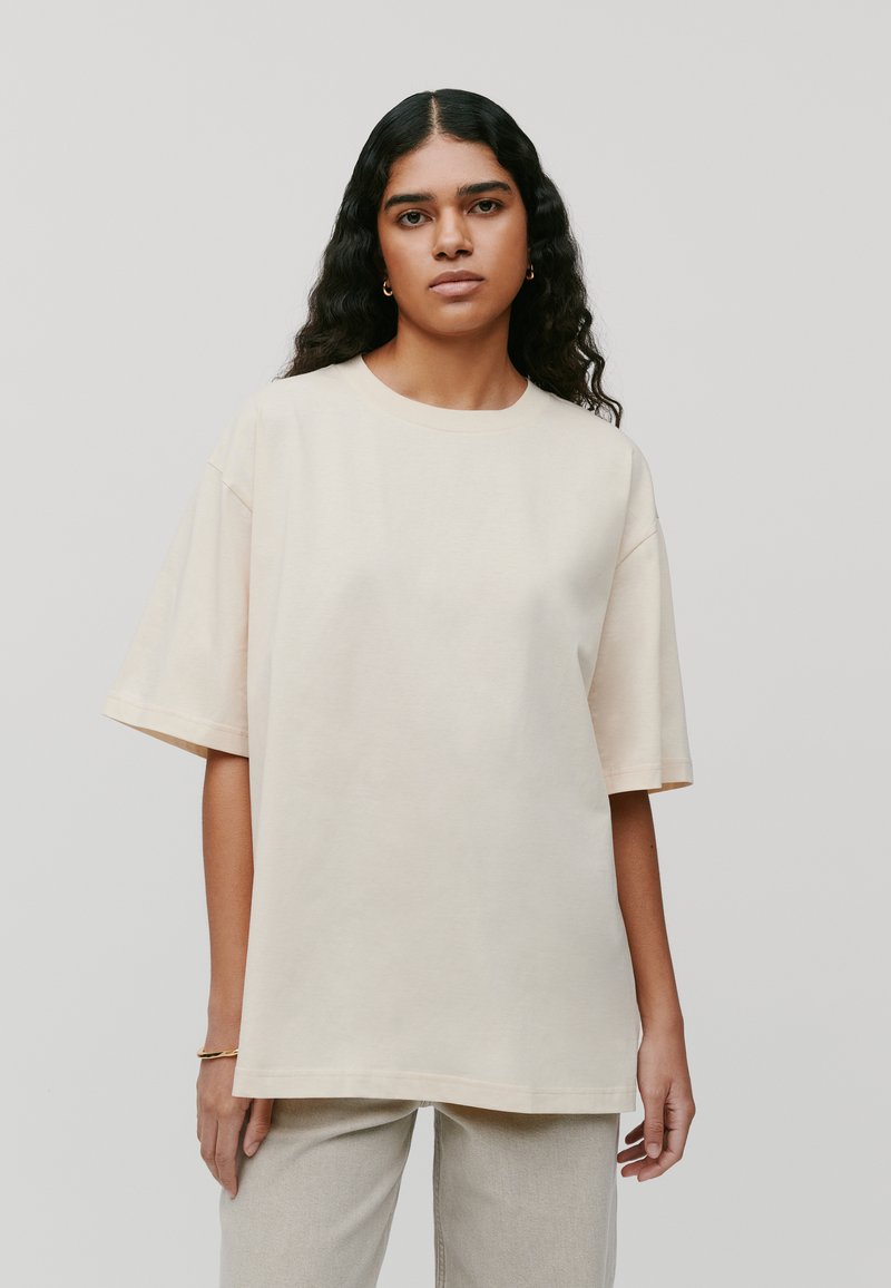 Przestronna koszulka w kolorze kremowym z krótkimi rękawami, okrągłym dekoltem i miękką bawełnianą fakturą. Wewnętrzne szwy widoczne wzdłuż dołu i rękawów.