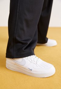 Vita lädersneakers med perforerat mönster, som har en ljusblå swoosh, matchade med svarta meshbyxor mot en gul golvyta.