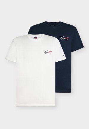 Fiatal férfi göndör hajjal, fehér Tommy Jeans pólót, bő kék farmert, zöld tornacipőt és karkötőt visel, egyszerű háttér előtt áll.