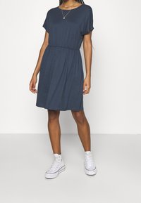 Marineblaue Kurzarmkleid mit gerafftem Taillenbereich, knielangem Saum und weicher Stoffqualität. Getragen mit weißen High-Top-Sneakern.