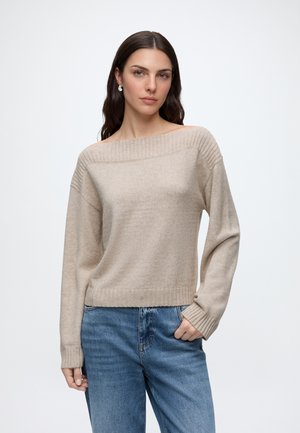 Femme aux longs cheveux foncés portant un pull beige épaules dénudées et un jean bleu, debout devant un fond clair uni.