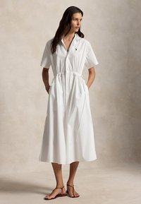 Femme portant une robe chemise mi-longue blanche à manches courtes avec taille à cordon et sandales, debout les mains dans les poches devant un arrière-plan neutre.