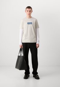 Lys beige t-skjorte med blå grafisk logo, lagdelt over en hvit langermet skjorte. Svarte slim-fit bukser og en svart totebag. Mørke sko.