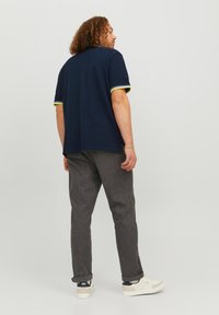 Marineblauwe shirt met korte mouwen en gele accenten op de manchetten, gecombineerd met grijze broek, gedragen met witte sneakers; subtiele textuur en een relaxte pasvorm.