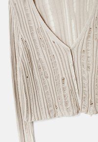 Lichtbeige gebreide cardigan met een diepe V-hals, knoopsluiting, verticale ribbeltextuur en rafelige randen voor extra detail.