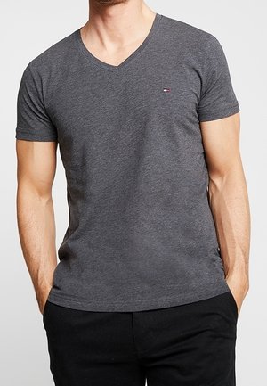 Mężczyzna noszący dopasowany, ciemnoszary T-shirt z dekoltem w serek i małym logo na piersi oraz czarne spodnie, ręce w kieszeniach.