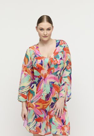 Vrouw met een lichte huid en haar naar achteren gebonden, gekleed in een kleurrijke bloemenjurk met lange mouwen en een V-hals, staand tegen een effen achtergrond.