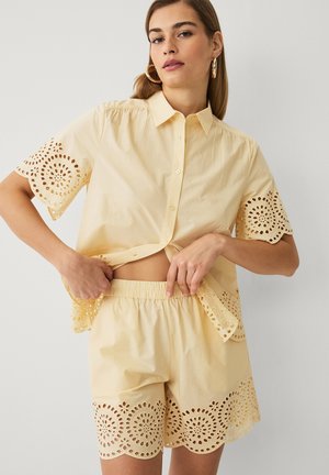 Kvinde iført en lysegul skjorte og matchende shorts med cirkulært udskåret blonde detaljer på ærmer, kant og shorts.